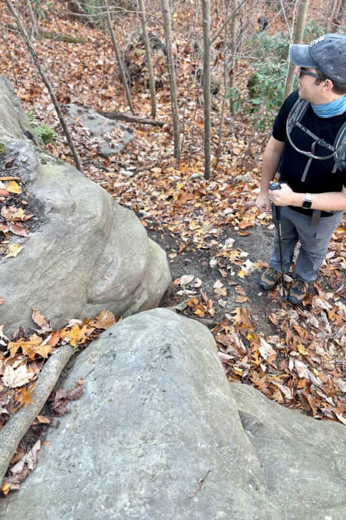 Hiker with trekking pole paused next to trailside boulders.