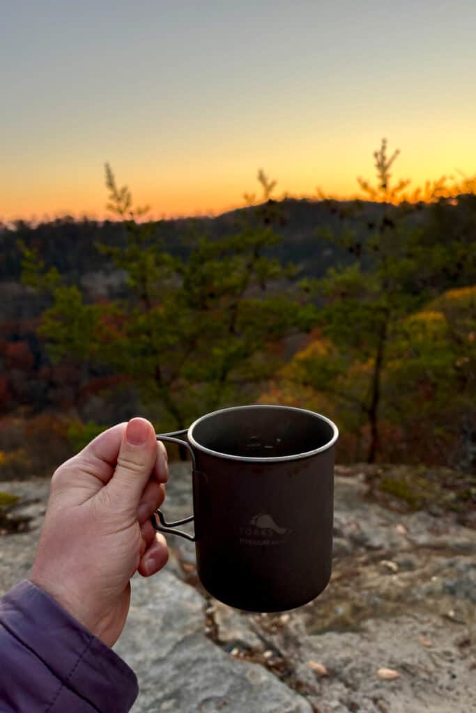 Hand holding titanium mug with sunset in background.