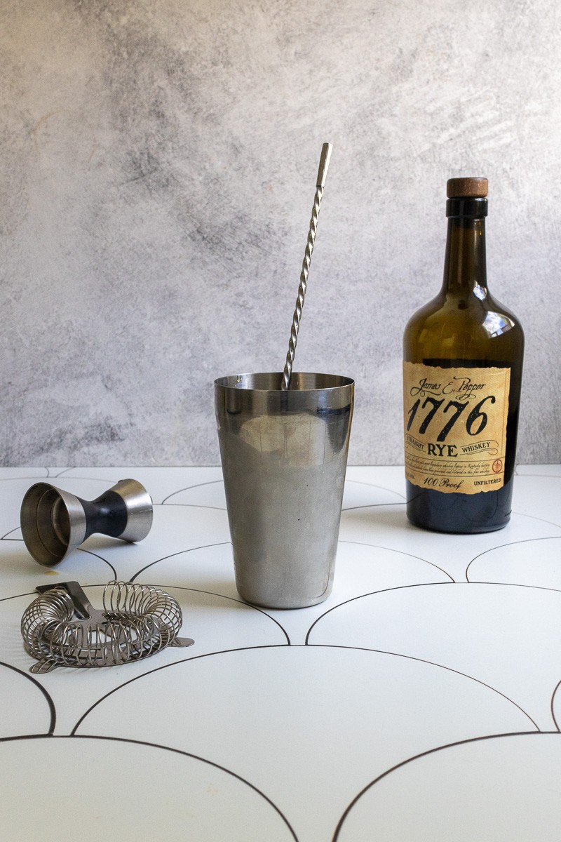 Rye whiskey, cocktail shaker with stir stick, jigger and strainer on a tile surface.