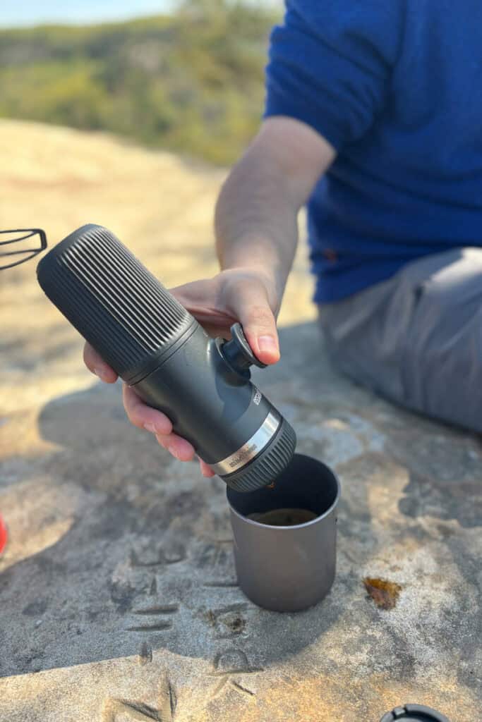Backpacking-friendly espresso maker held over mug by hiker seated on rocky surface.