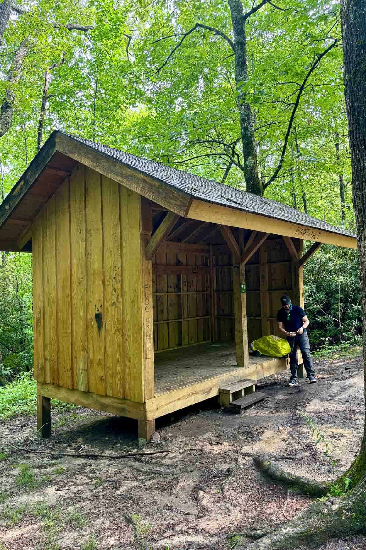 Hiker adjusting backpack at small wooden trail shelter along Art Loeb Trail.