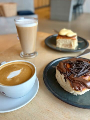 Flat white in coffee cup next to sweet roll topped with chocolate.