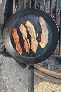 Four slices of bacon being cooked in cast-iron pan over campfire.