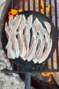 Slices of bacon cooking in cast-iron skillet over campfire.