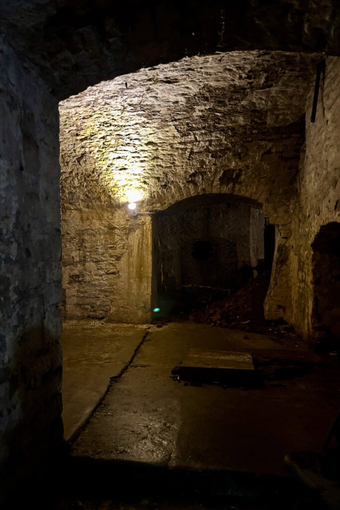 Part of a Cincinnati underground brewery tour, a stone-lined room with damp floor and small alcoves through arched doorways, lit with one electric light.