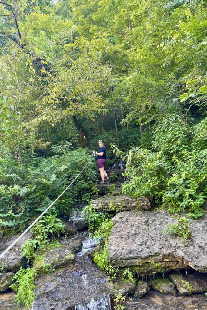 Woman in shorts and t-shirt holding onto rope while ascending slippery rock trail.