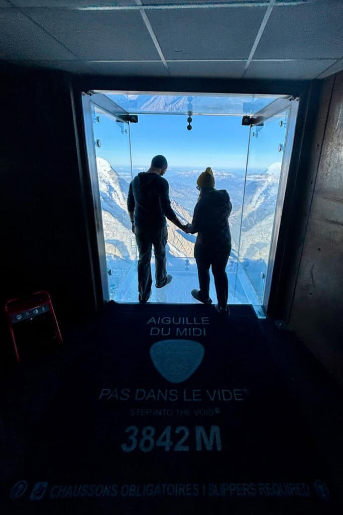 Man and woman preparing to step out on transparent viewing platform atop Aiguille du Midi summit.