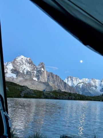 View from tent entrance across lake to snow-covered mountains with moon in the dark blue sky.