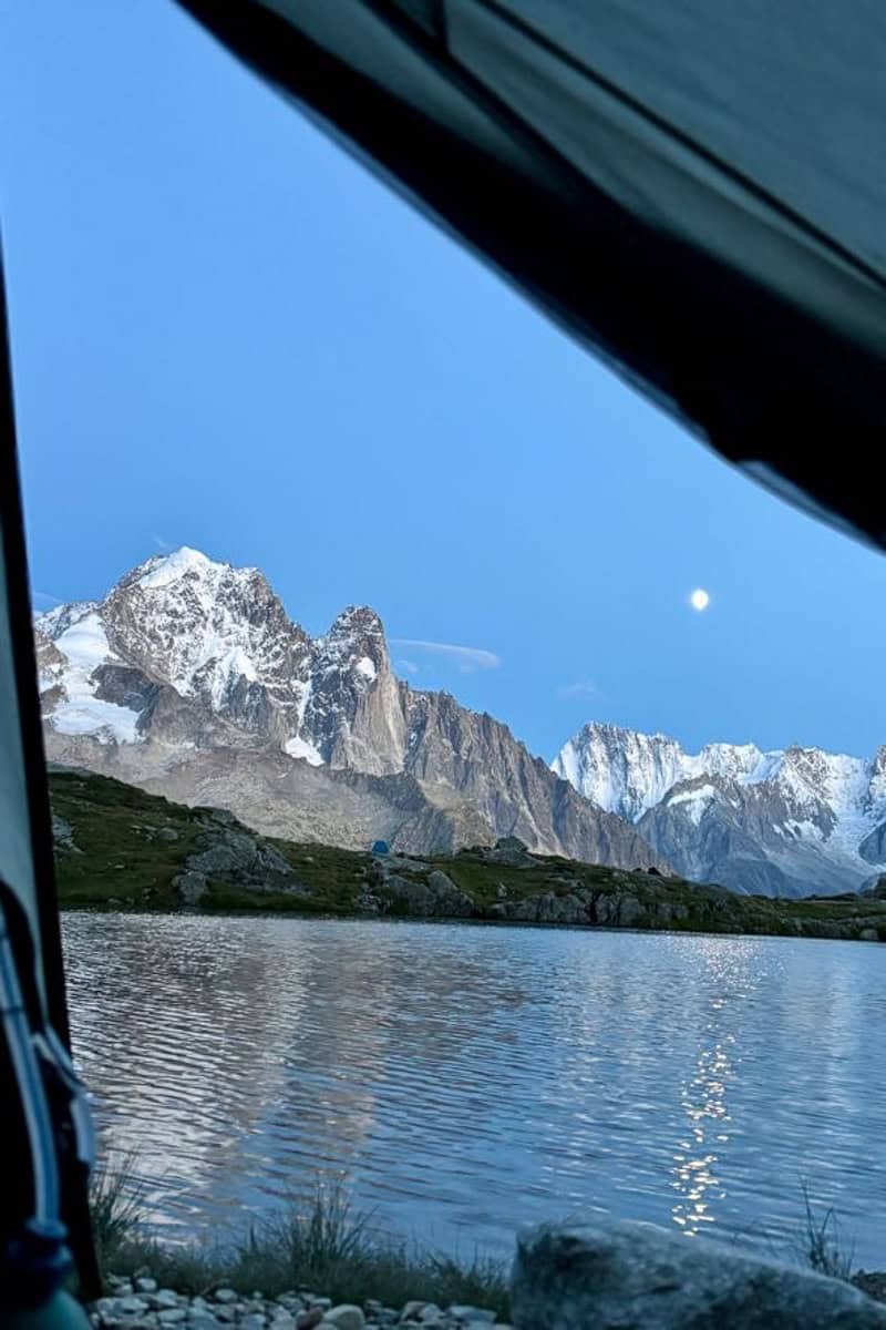 View from tent entrance across lake to snow-covered mountains with moon in the dark blue sky, seen while camping the Tour du Mont Blanc.