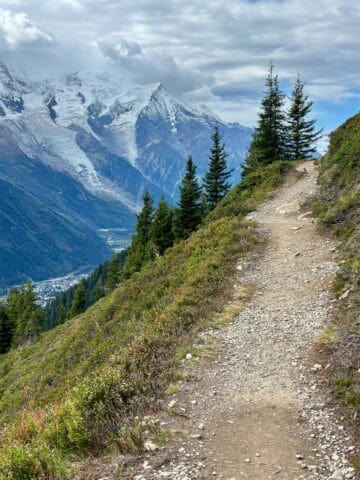Dirt trail ascending mountainside with view of snow-covered peaks across valley on the Tour du Mont Blanc.