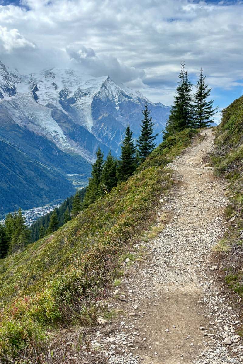 Dirt trail ascending mountainside with view of snow-covered peaks across valley on the Tour du Mont Blanc.