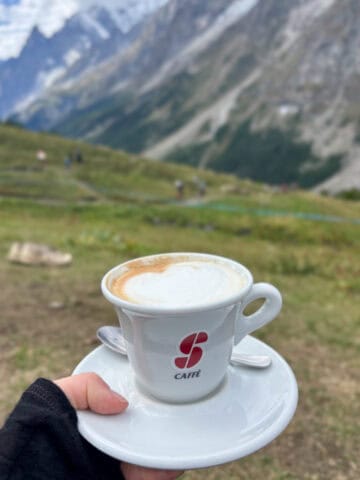 Ceramic cup of coffee and saucer with mountain view in background.