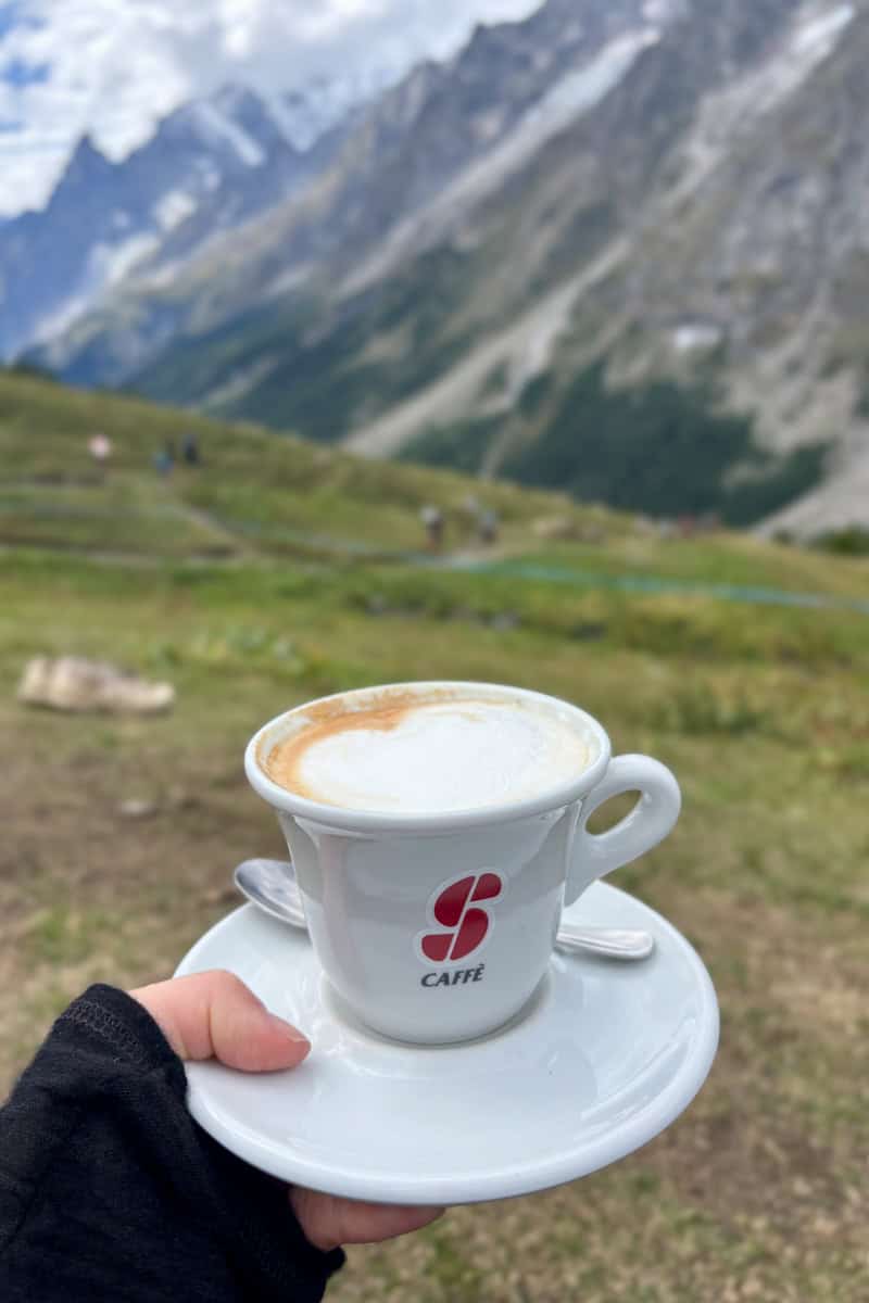 Ceramic cup of coffee and saucer with mountain view in background.