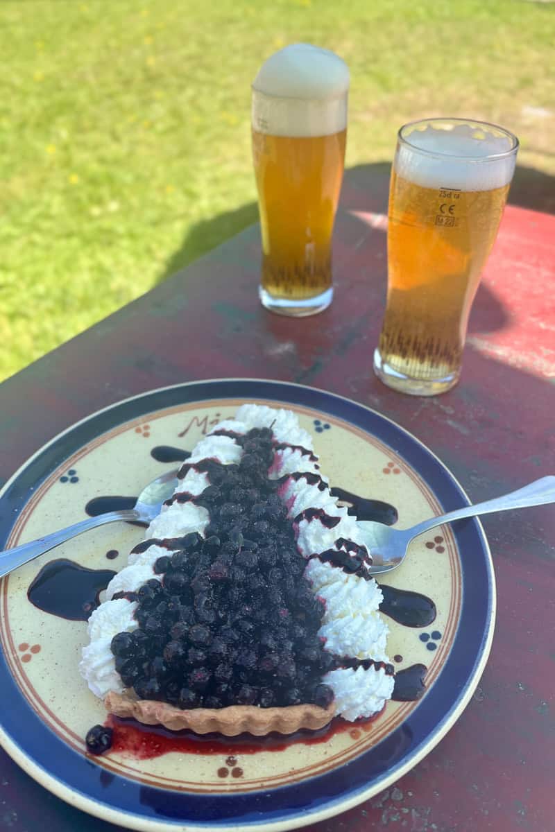 Slice of blueberry pie and whipped cream on plate with glasses of beer in background, options among the best food and drink on the Tour du Mont Blanc.