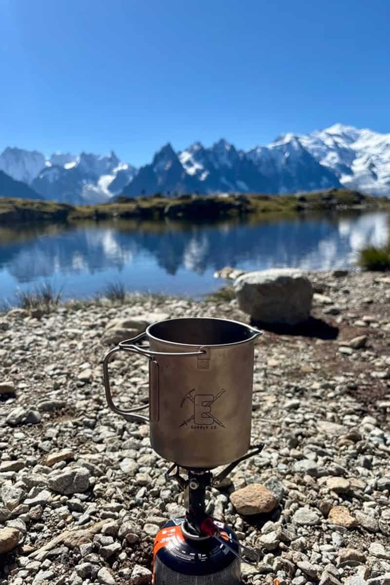 Cook pot attached to small fuel canister on gravel surface with mountains and lake in background.