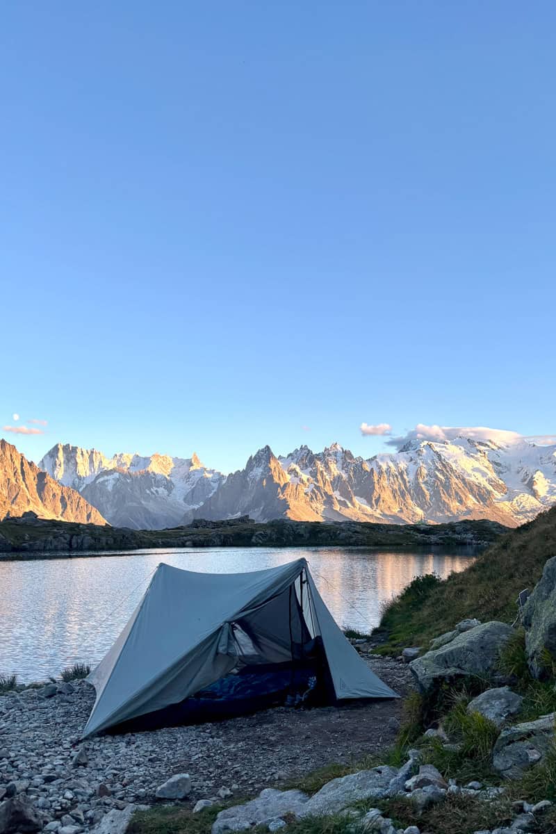Backpackers tent pitched at edge of lake with snow-covered mountains across the water.