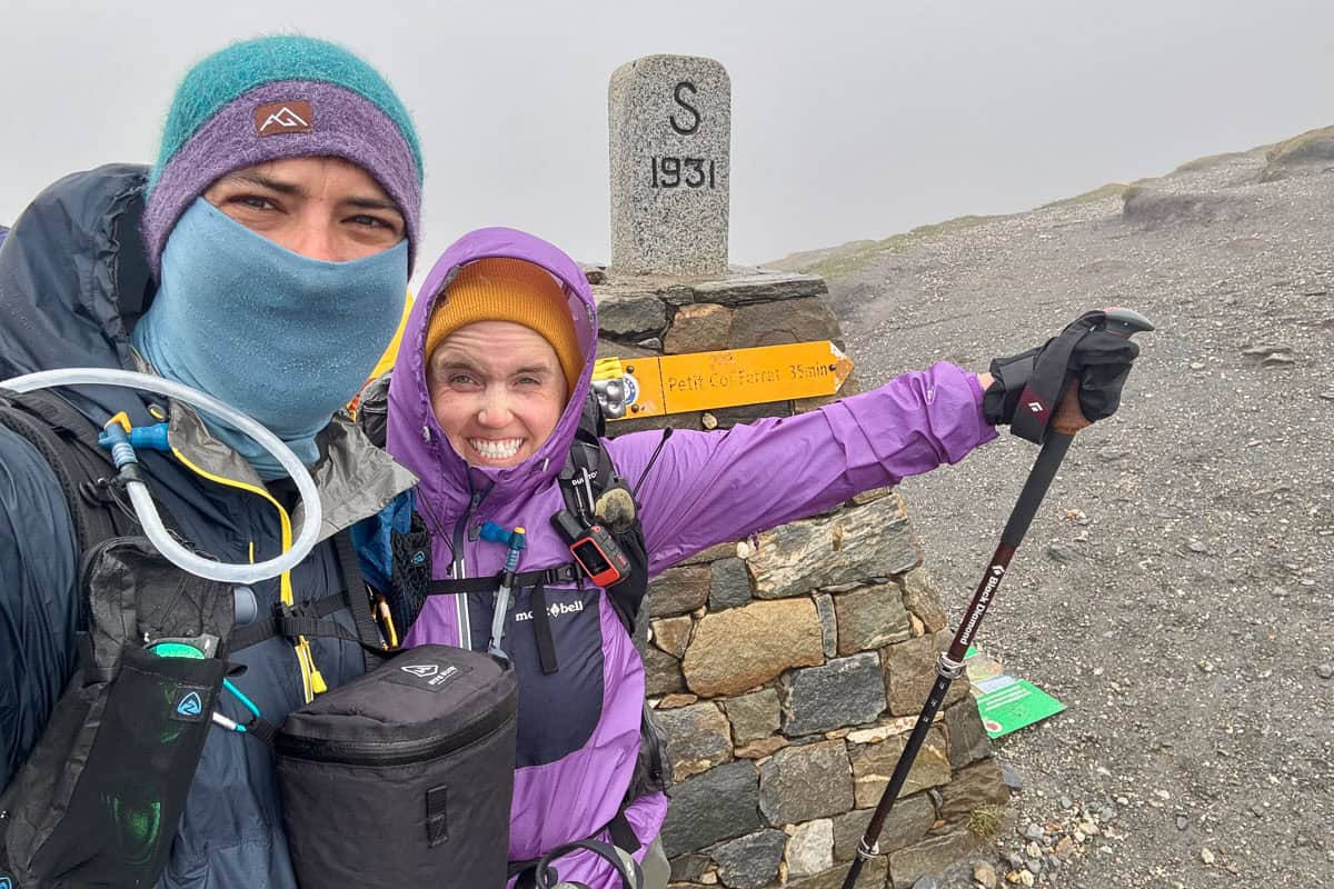 Two hikers bundled up for cold weather on foggy mountainside next to trail marker on the Tour du Mont Blanc.