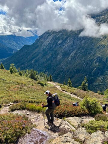 Woman carrying backpack and trekking pole hiking down hill with cloud-covered mountains in view.