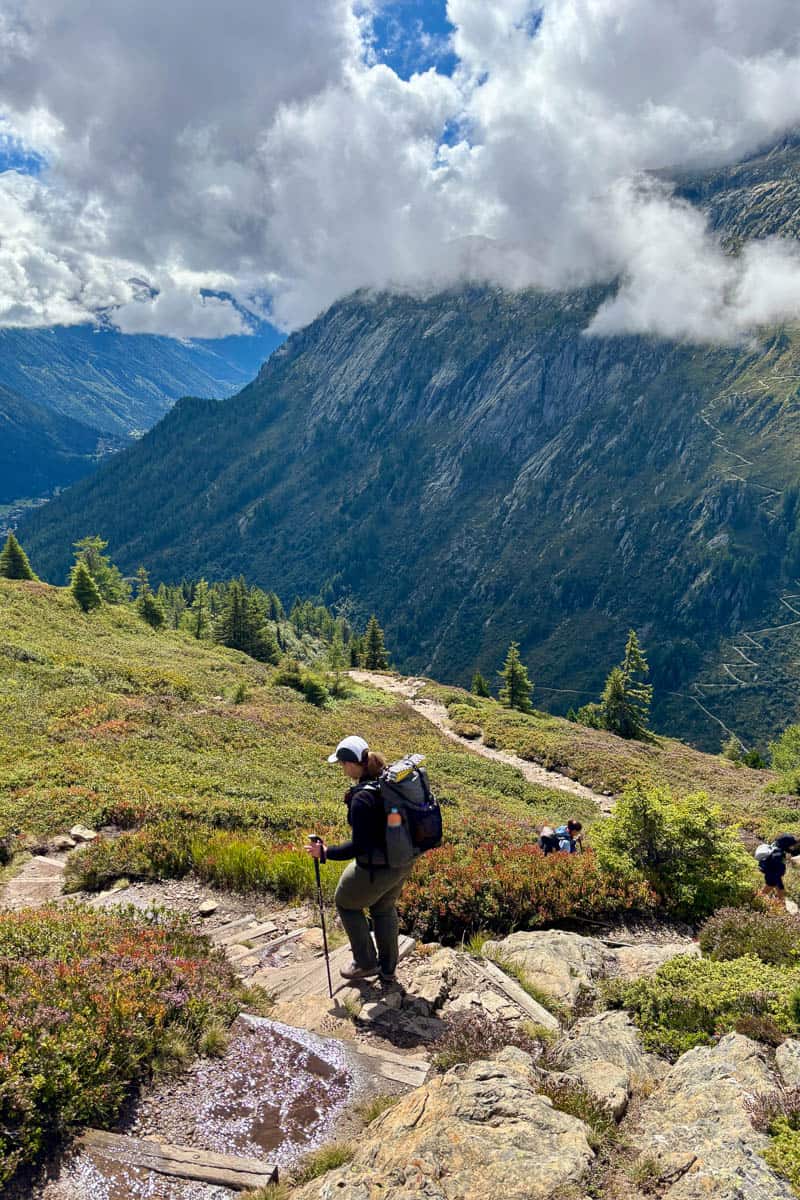 Woman carrying backpack and trekking pole hiking down hill with cloud-covered mountains in view.