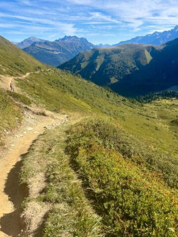 Dirt trail along grassy mountainside with peaks in the distance.