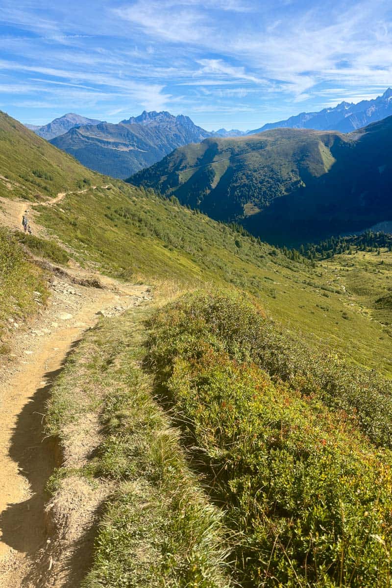 Dirt trail along grassy mountainside with peaks in the distance.