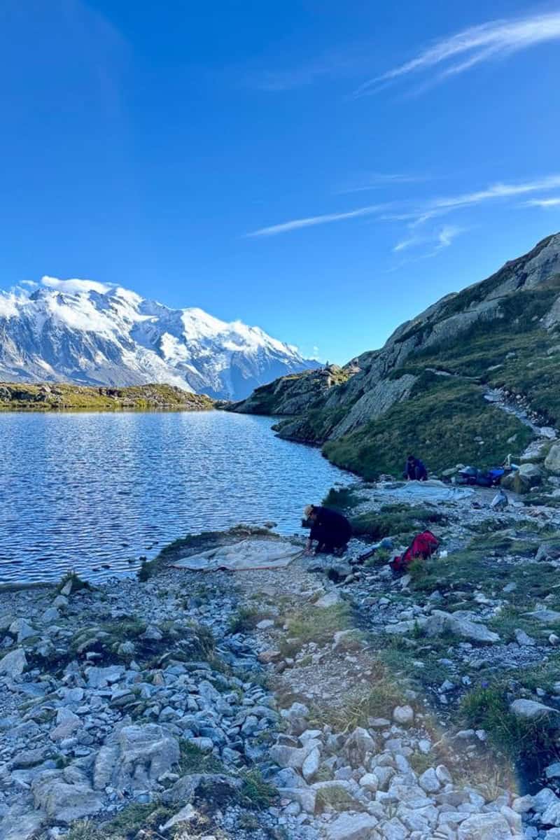 Campers pitching tents on rocky lakeshore with view of snowy mountain range.