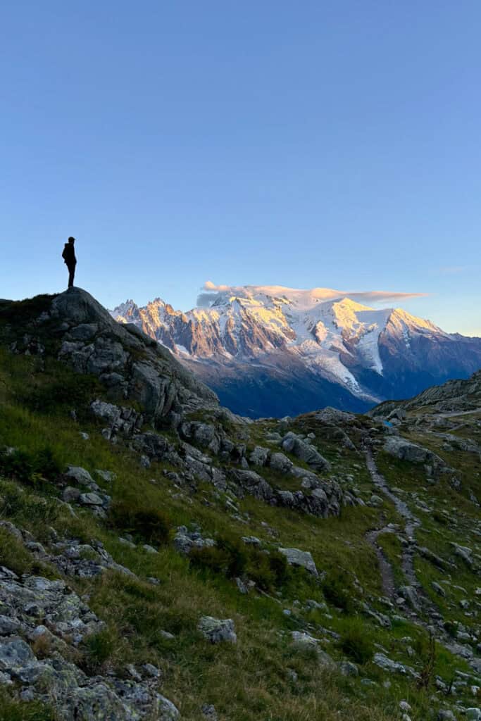 Single hiker in silhouette standing at top of rocky outcrop with view of snowy mountains on Tour du Mont Blanc stage 10.