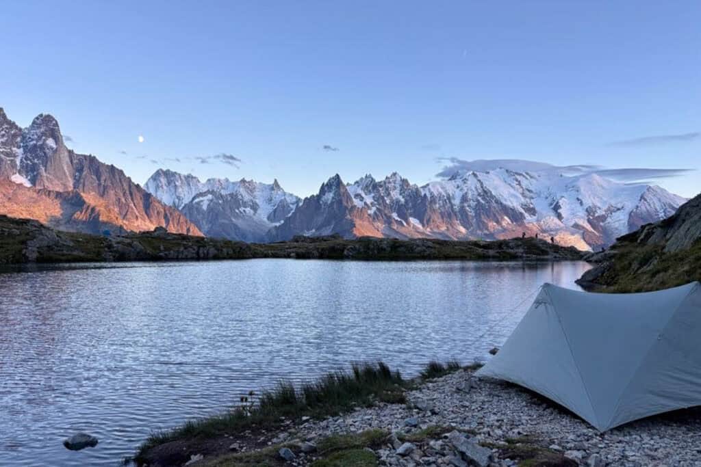 Tent pitched at edge of lake with view across lake of snowy mountain range in fading evening light.