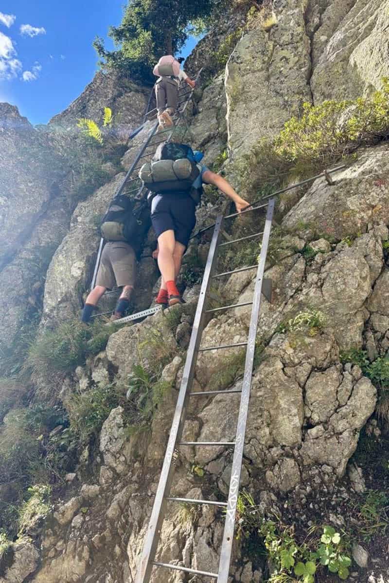 View from below of hikers climbing ladders leaned against cliff face.