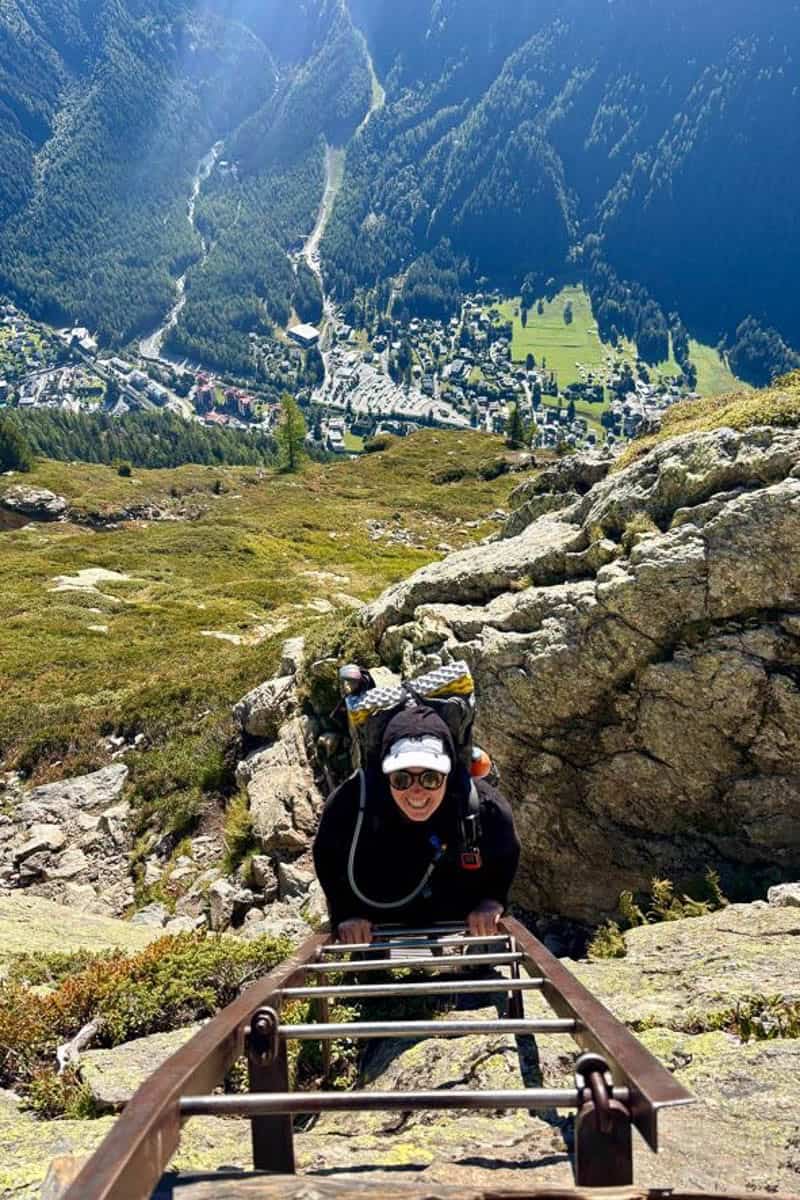 View from above of smiling hiker climbing metal ladder with Alpine valley below on Tour du Mont Blanc stage 10.