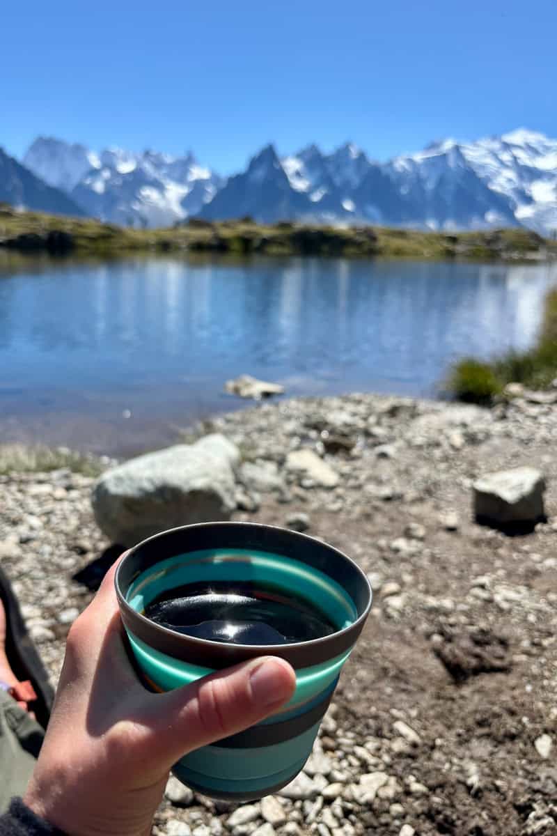 Collapsible cup containing coffee with Alpine lake and mountain view in background.