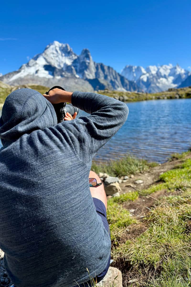 Hiker wearing hoodie using camera to photograph lake and snow-covered mountain peaks.
