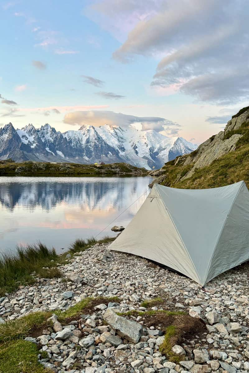 Tent pitched at edge of lake with view of snowy mountain range in early morning light.