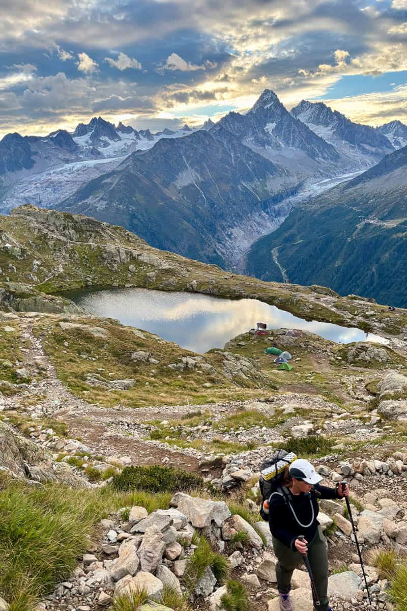 Hiker ascending steep rocky trail with snowy mountain range and alpine lake in background on Stages 10 and 11 of the Tour du Mont Blanc.