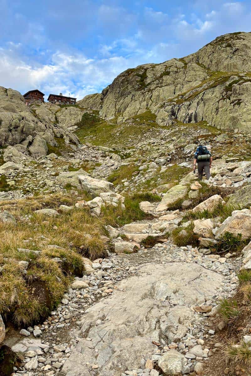 Hiker ascending rocky mountainside with hikers refuge just visible at top of ridge.