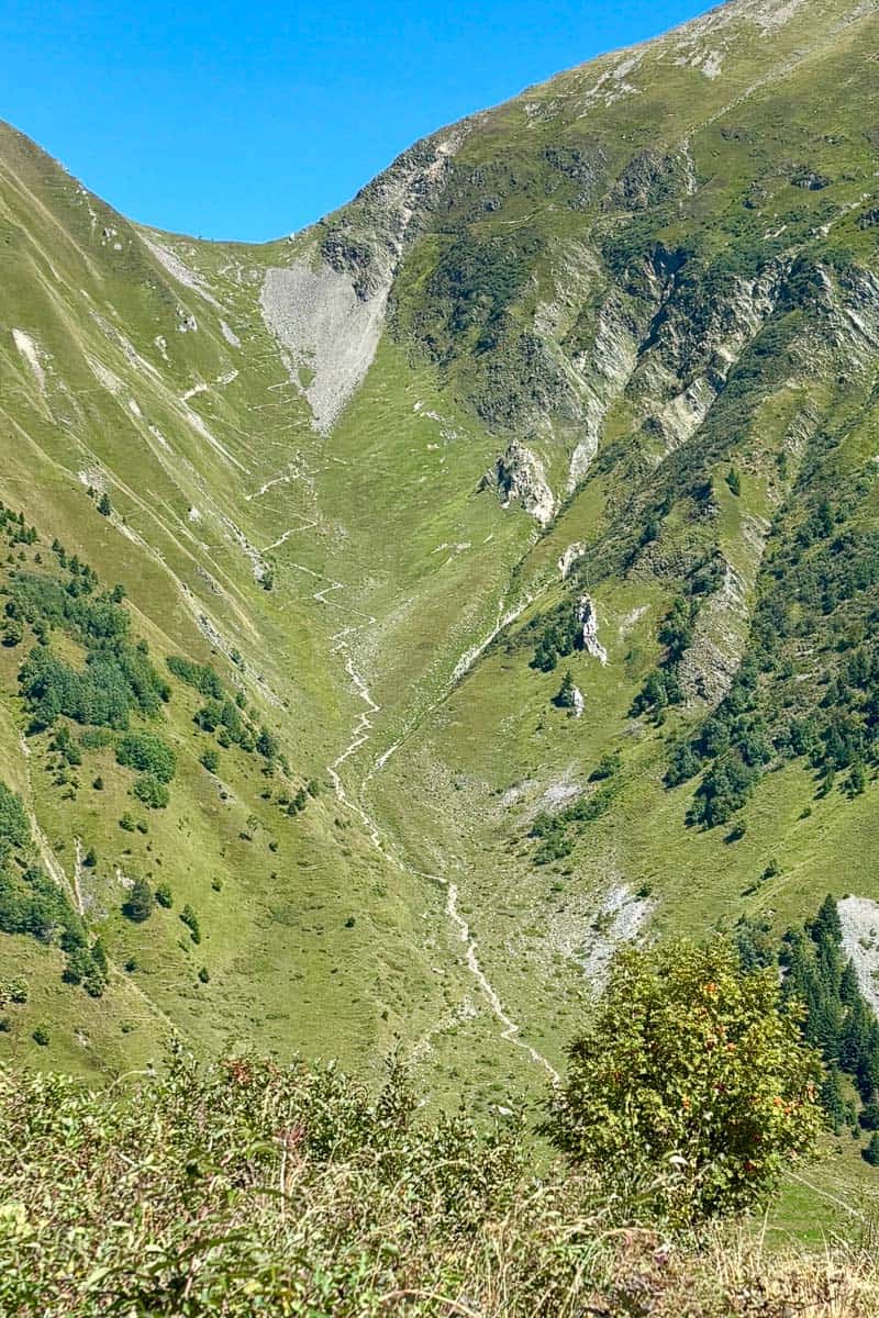 Steep zig-zagging trail visible down grassy mountainside.