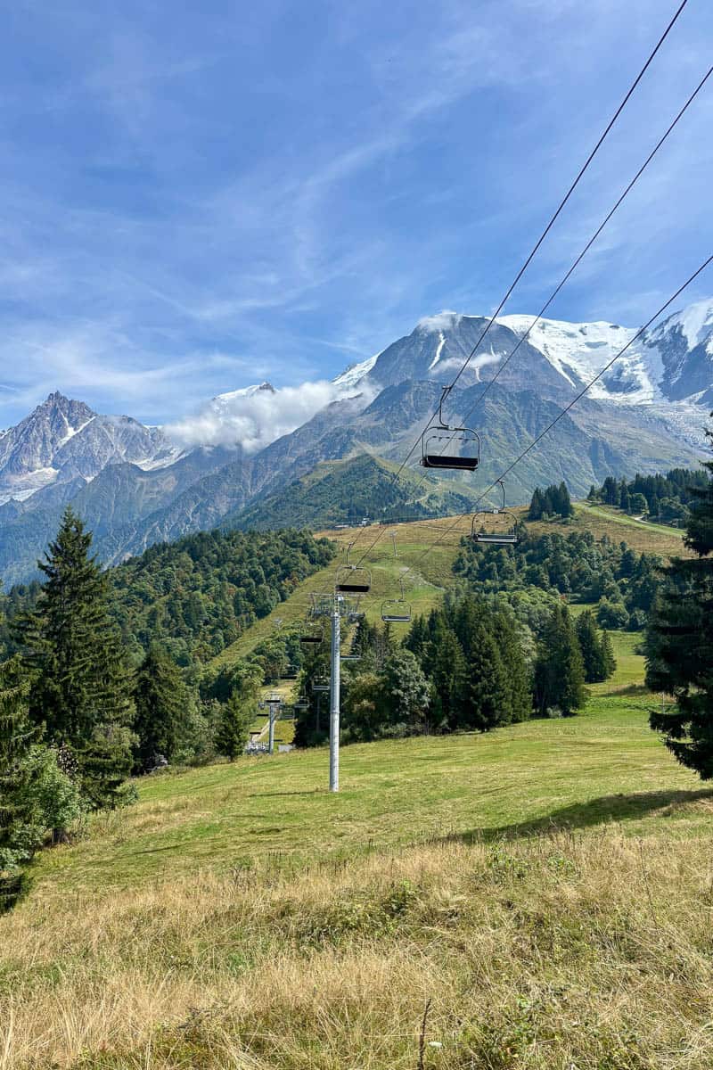 Chair lift above grassy mountainside with snow-covered peaks in distance.