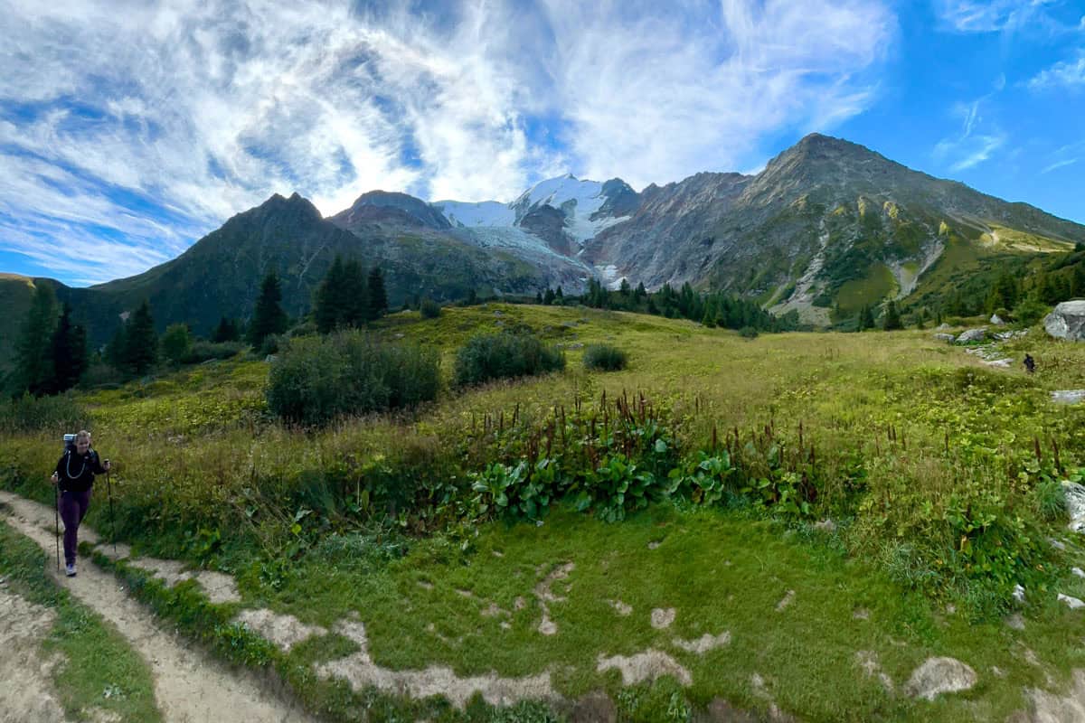 Backpacker on dirt trail alongside Alpine meadow and mountain peak.