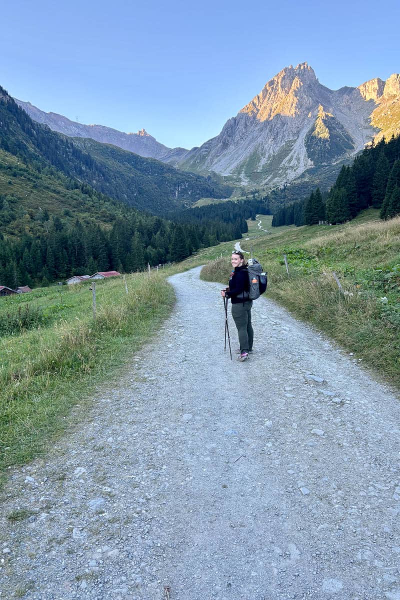 Backpacker on gravel trail with jagged mountain peak illuminated by sunlight in the background.