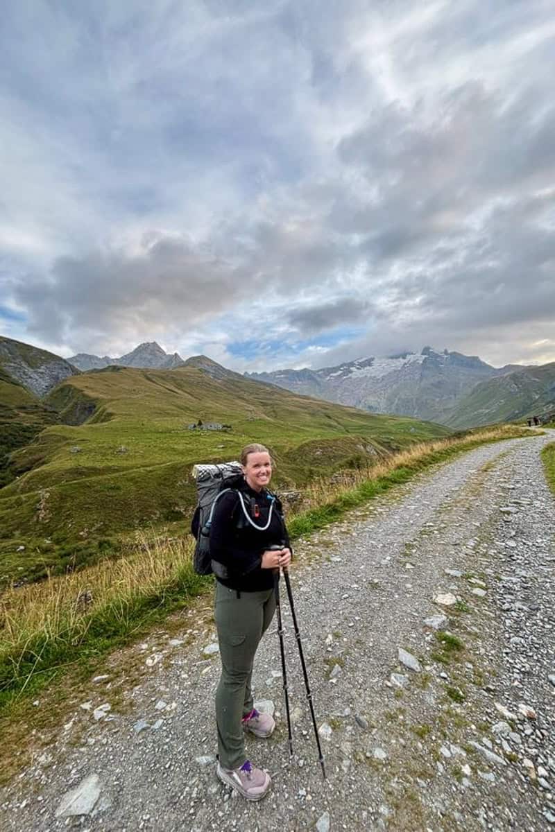 Woman carrying backpack and trekking poles on gravel trail with mountains in distance.