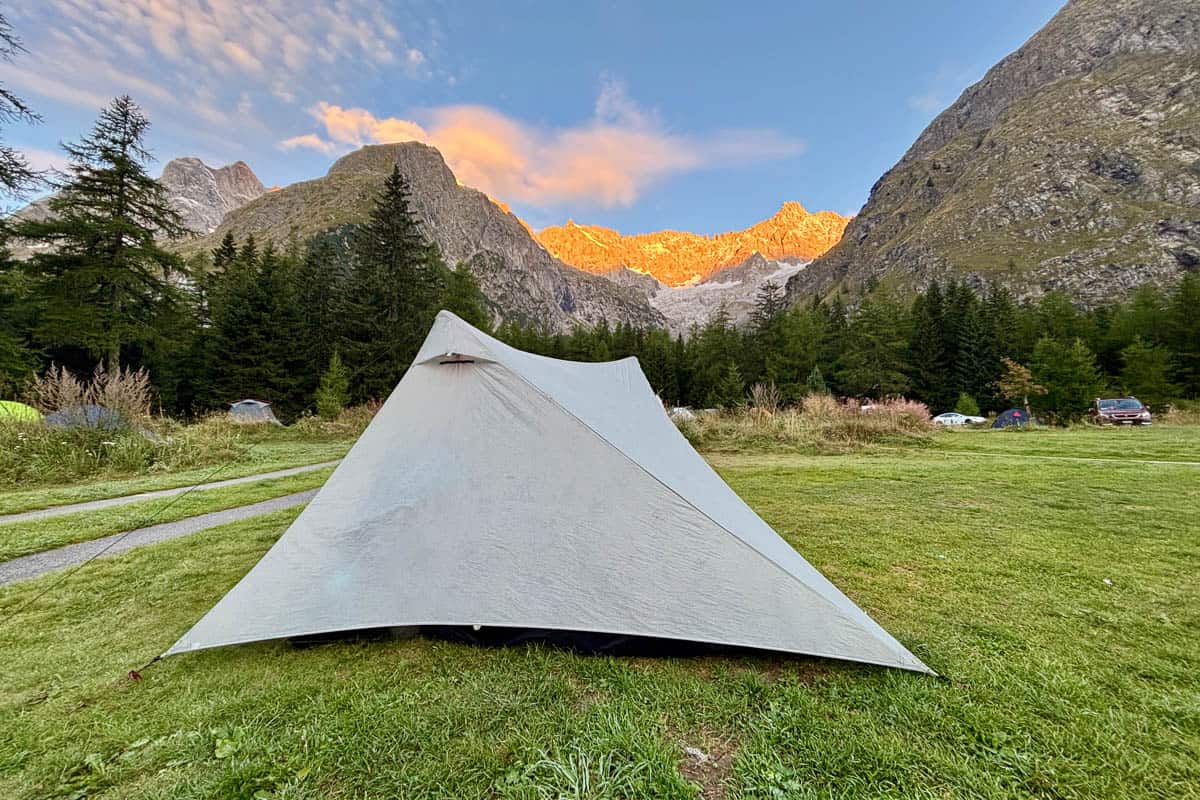 Backpackers tent pitched in grassy field with view of sun illuminating distant rugged peaks.