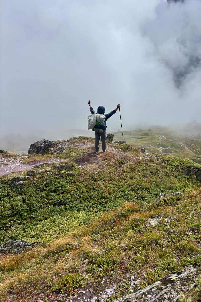 Backpacker with arms raised triumphantly toward foggy view on Tour du Mont Blanc Stage 9.