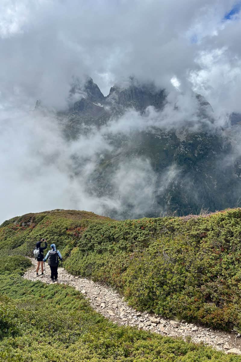 Hikers descend rocky trail on Tour du Mont Blanc Stage 9 with jagged mountain peaks barely visible through gaps in clouds.