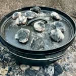 Dutch oven covered by lid and set over hot charcoal to bake, with several briquettes placed on top of the lid.