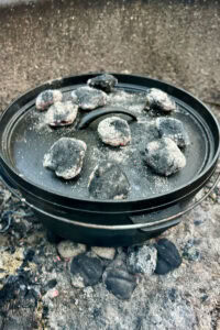 Dutch oven covered by lid and set over hot charcoal to bake, with several briquettes placed on top of the lid.