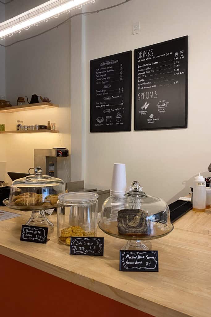 Food and drink menu board on wall above ordering counter, with display cases of baked goods at Floating Lotus Kitchen.