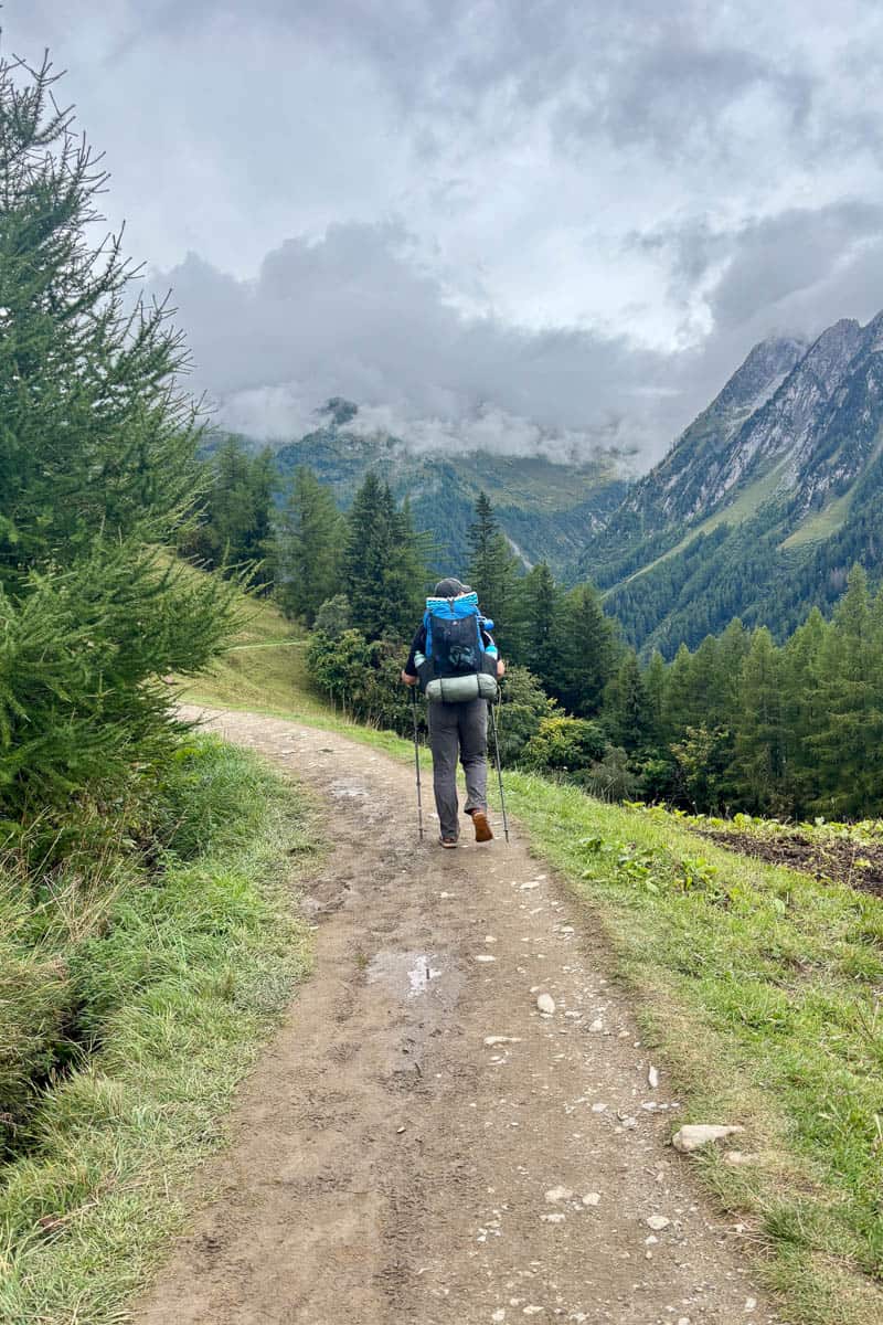 Hiker on dirt path viewing cloud-obscured alpine mountainsides across the valley on Stage 8 of the Tour du Mont Blanc.