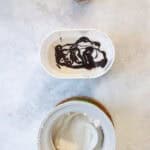 Ice cream with ladle in ice cream maker basin, next to container partially filled with bourbon ice cream and layer of chocolate sauce.