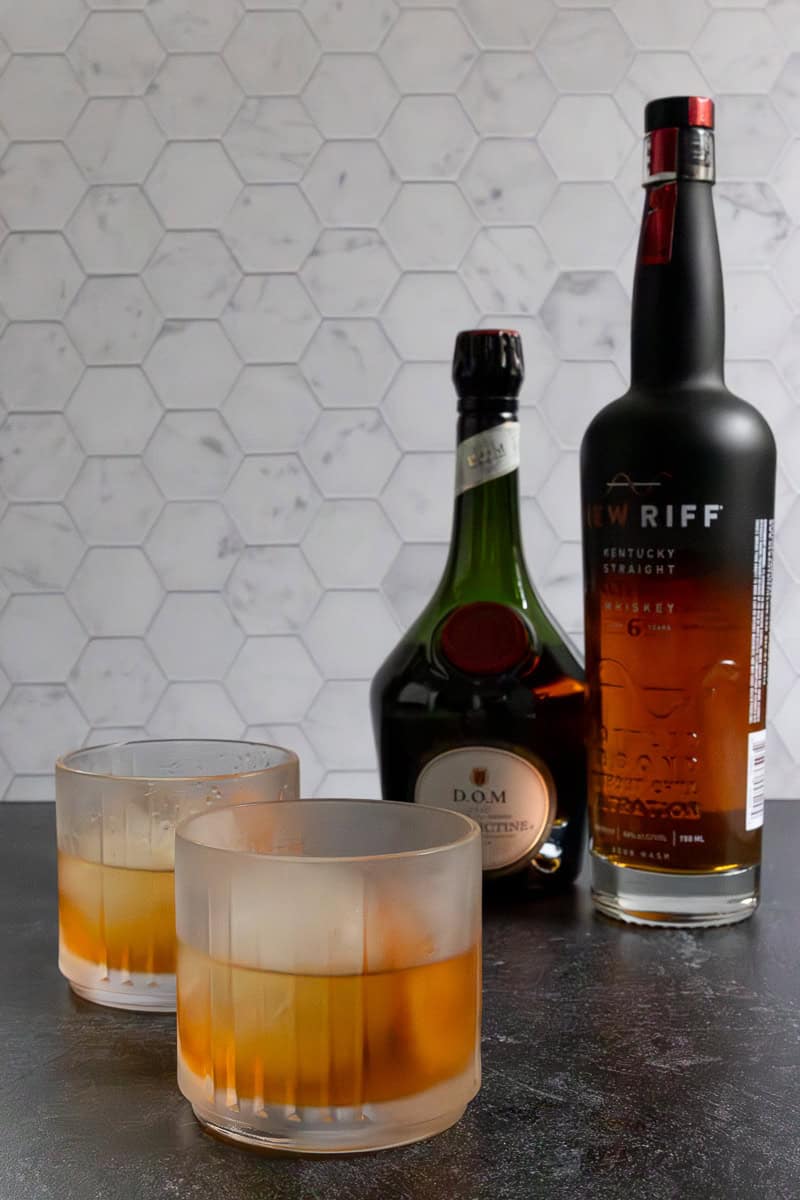 Two Monte carlo cocktails in glasses next to bottles of Benedictine and whiskey.