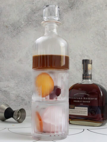Two chilled rocks glasses stacked below small pitcher containing old fashioned cocktail, with bottle of bourbon and cocktail jigger in background.